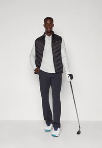 Colete acolchoado preto sobre uma camisa polo cinza clara, calças escuras e sapatos de golfe brancos. Segurando um taco de golfe, em pé contra um fundo simples.