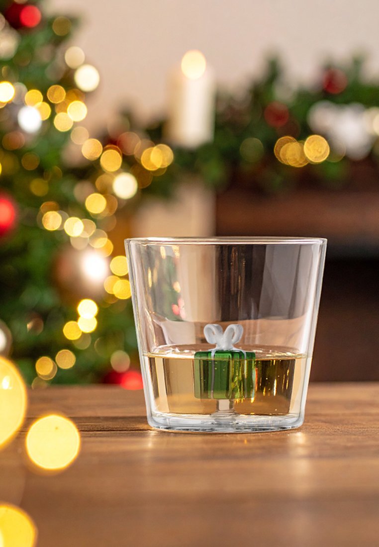 Przezroczysty szklany tumbler wypełniony jasnym płynem, z małym zielonym pudełkiem prezentowym z białą wstążką na dole. Udekorowane tło.