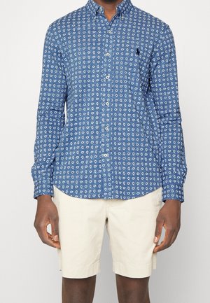 Homme portant une chemise bleue à manches longues avec des motifs blancs et un short beige arrivant au genou.