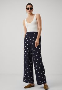 Marineblå polkadottbukser med vid benvidde og glat tekstur samt hvide cirkulære mønstre, sammen med en tætsiddende hvid top og brune mules.