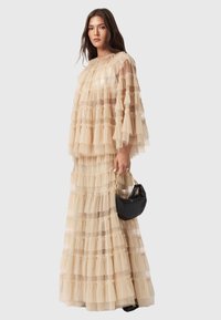 Μπεζ τούλινο φόρεμα με στρώσεις φρουφρού, με στρογγυλή λαιμόκοψη και φαρδιά μανίκια. Συνδυ�ασμένο με μια μαύρη τσάντα.