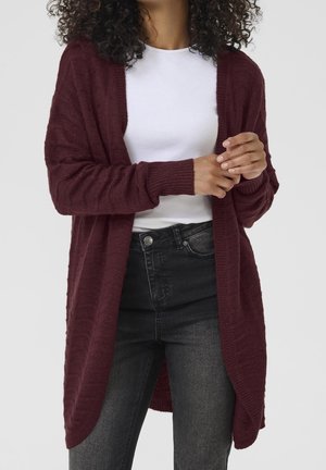 Kobieta w teksturowanym bordowym kardiganie, białej prążkowanej koszulce i czarnych dżinsach, stojąca z złączonymi dłońmi przed sobą.