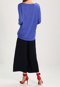 Blå blus med prickar och långa ärmar, med en vid halsringning, i kombination med vida svarta byxor och röda högklackade skor med ankelremmar.