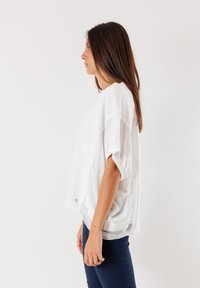 Maglietta bianca oversize a maniche corte con scollatura rotonda, spacchi laterali e una texture liscia, abbinata a pantaloni aderenti blu scuro.