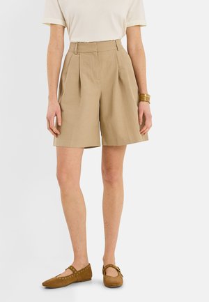 Mujer que lleva pantalones cortos plisados beige de cintura alta, camiseta blanca de manga corta, zapatos planos marrones con tachuelas doradas y una pulsera dorada, de pie.