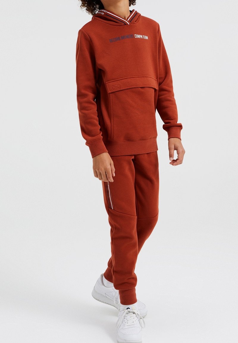 Röd sweatshirt med huva och ficka fram, matchande byxor. Har randiga detaljer på kragen och sidan av byxorna.