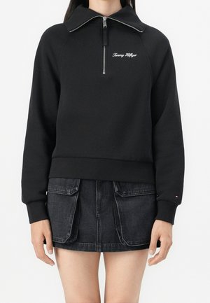 Femme portant un sweat-shirt noir Tommy Hilfiger à zip quart avec manches longues et une mini-jupe en denim noire avec poches avant.