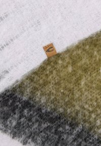 Wollen dekenmix met zachte texturen in wit, olijfgroen en grijs, voorzien van een klein leren label aan de rand.