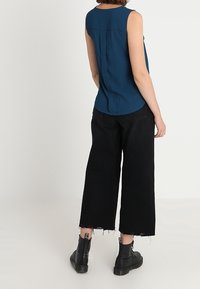 Femme portant un top sans manches de couleur teal foncé, un jean court à jambes larges noirs avec des ourlets effilochés et des bottines noires, se tenant contre un fond blanc.