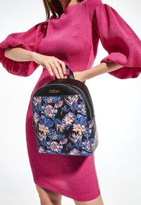 Žena v jasně růžových šatech s nabíranými rukávy drží černý batoh s modro-růžovým květinovým vzorem.