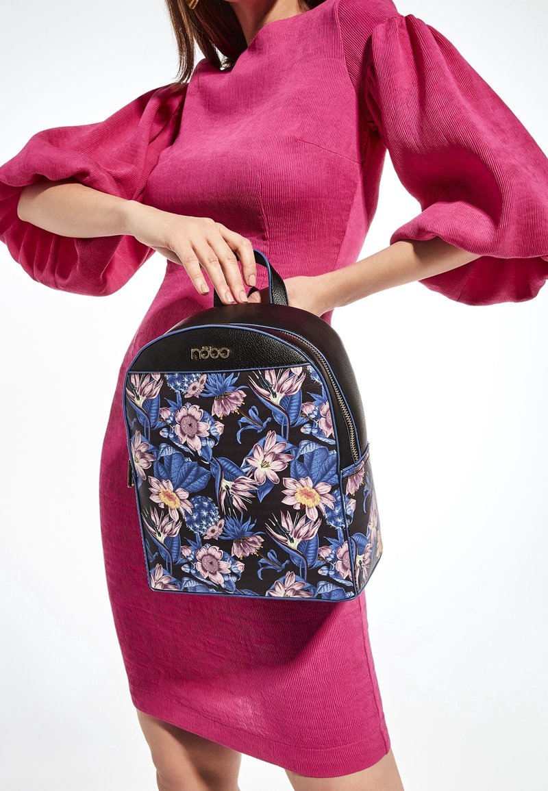 Žena v jasně růžových šatech s nabíranými rukávy drží černý batoh s modro-růžovým květinovým vzorem.