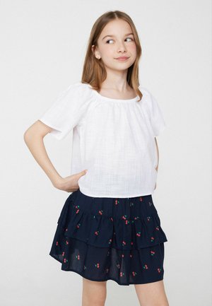 Fille aux cheveux châtain clair portant un chemisier blanc et une jupe bleu marine avec imprimé de cerises rouges, debout les mains sur les hanches, regardant sur le côté.