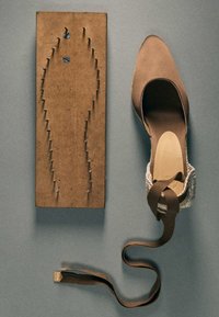 Chaussure à talon haut marron avec bride à la cheville à côté d’une planche en bois avec des clous disposés en contour de pied sur une surface grise.
