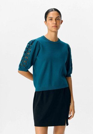 Maglione lavorato a maglia color turchese con maniche a sbuffo, con dettagli a taglio, abbinato a una gonna nera con uno spacco laterale. Tessuto liscio e design aderente.