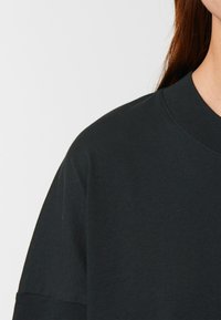 Juodas medvilnės džemperis su apvalia iškirpte ir trumpomis rankovėmis, pasižymintis lygiu paviršiumi ir laisvu siluetu, matomas dalis iškirpčių ir peties.