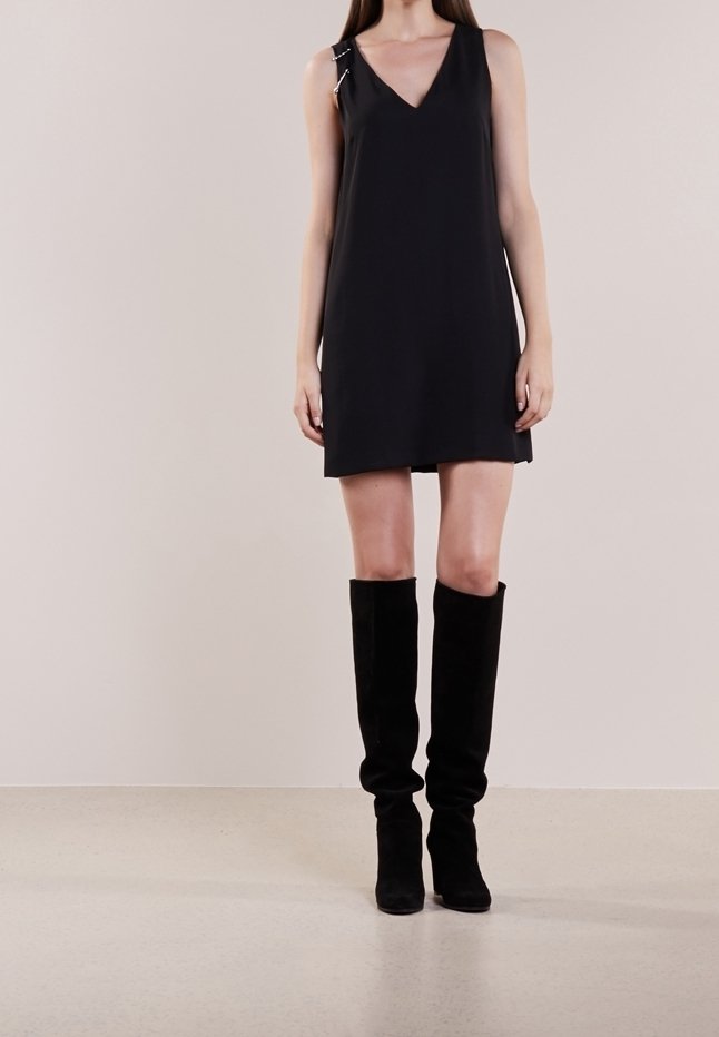 Czarna sukienka bez rękawów z dekoltem w serek, ozdobiona srebrną ozdobną agrafką na lewej ramieniu, zestawiona z czarnymi kozakami do kolan.