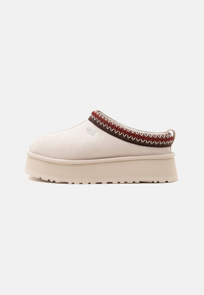 Chaussure beige à enfiler avec semelle épaisse, finition brodée marron et rouge autour de l'ouverture, et logo "UGG" sur le côté.