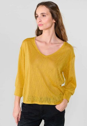 Haut léger à manches longues jaune en V, avec une coupe décontractée, une texture légèrement transparente et des poignets roulés, associé à un pantalon sombre.
