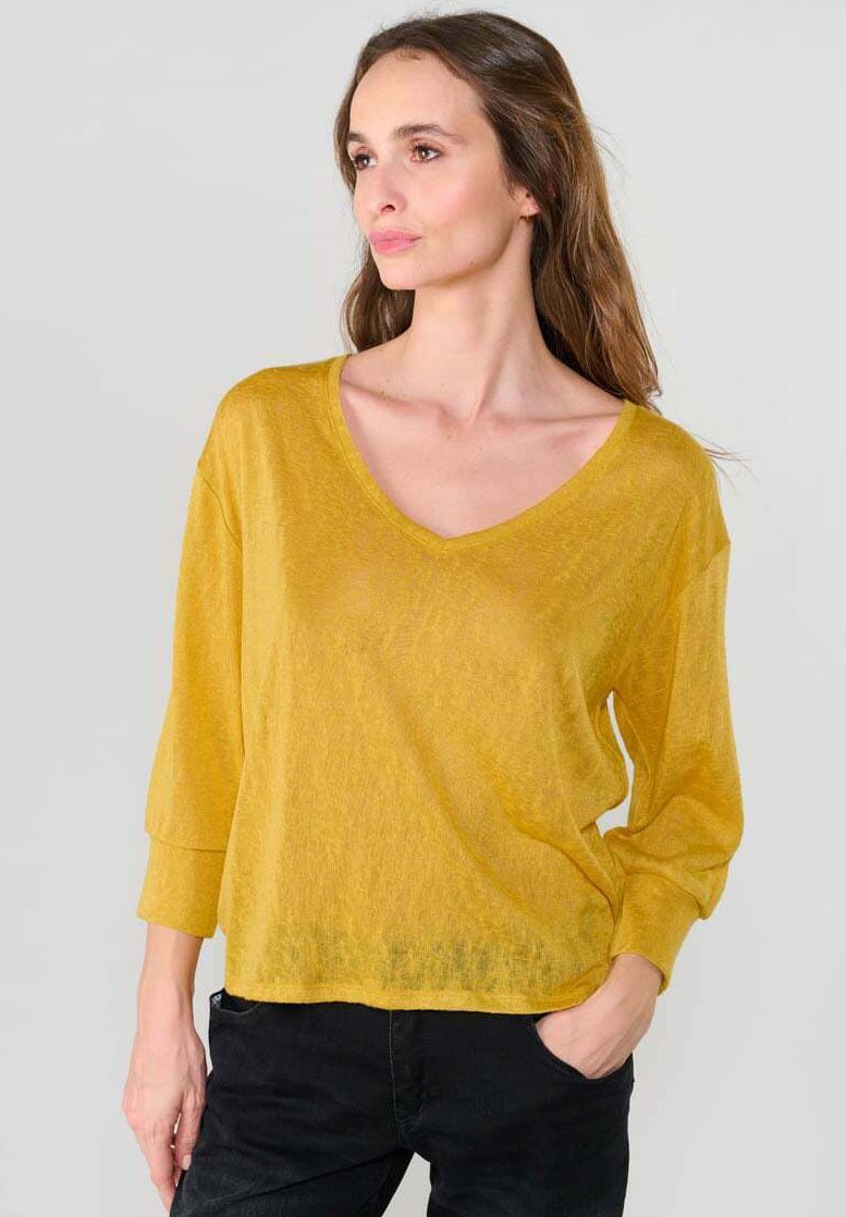 Haut léger à manches longues jaune en V, avec une coupe décontractée, une texture légèrement transparente et des poignets roulés, associé à un pantalon sombre.