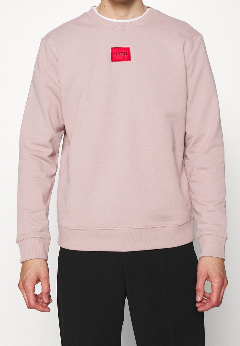 Personne portant un sweat-shirt rose clair avec un écusson rectangulaire rouge marqué "HUGO" sur la poitrine et un pantalon noir.