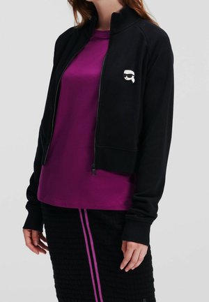 Kvinde iført en sort lynlåstrøje med en lille hvid ansigtspatch og en lilla skjorte under, kombineret med et sort skørt med pink striber.