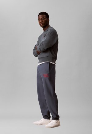 Mand stående med armene krydsede iført en grå sweatshirt, mørkegrå Calvin Klein joggingbukser og hvide sneakers på en ensfarvet baggrund.