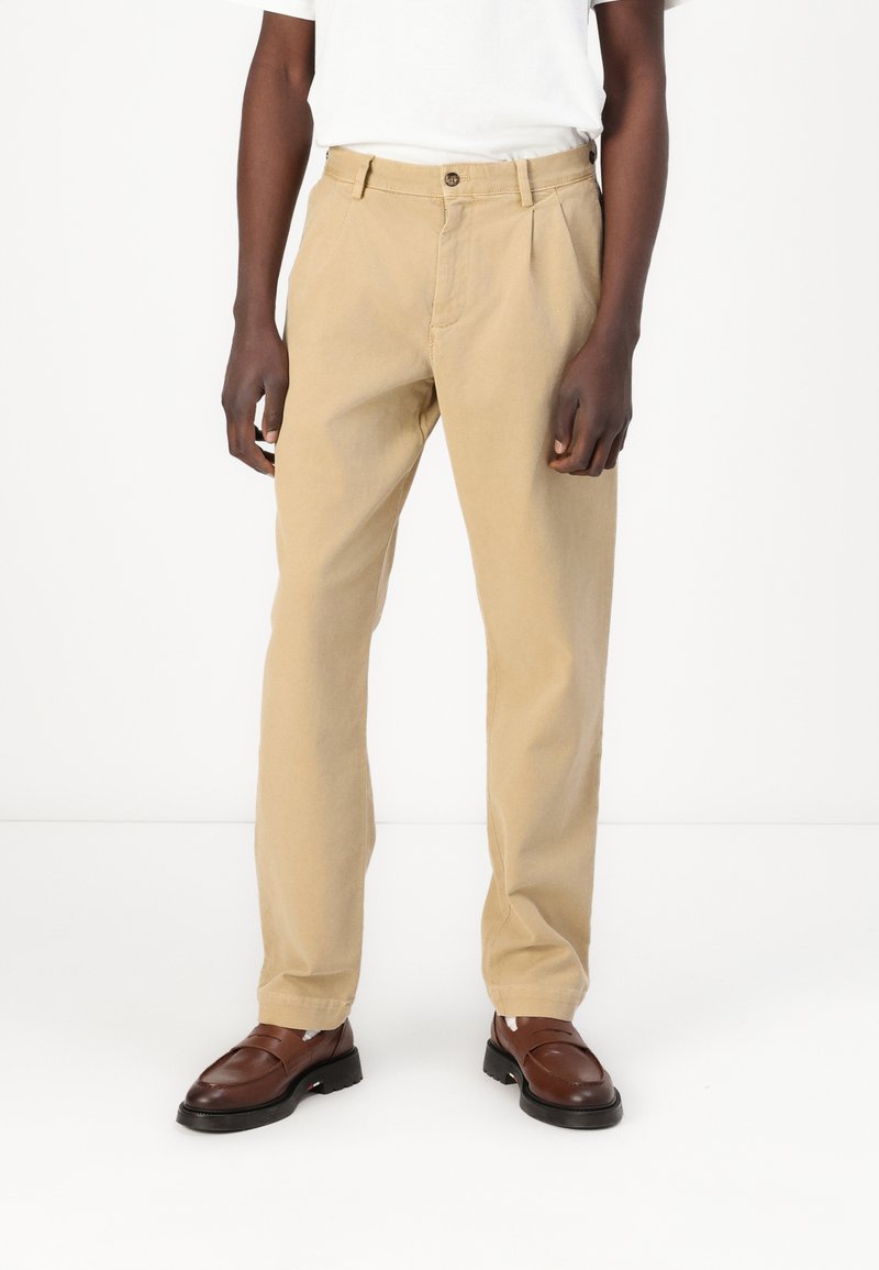 Beige bomullbukser med avslappet passform, frontfold og knappelukking. Brukt med brune slip-on sko. Jevn tekstur og klassisk design.