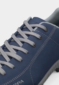 Μπλε casual αθλητικό παπούτσι με ανοιχτό γκρι κορδόνια, λευκή ραφή και υφασμένη γκρι σόλα, εμφανισμένο σε κοντινή λεπτομέρεια.