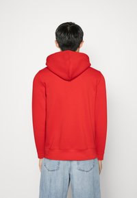 Punainen huppari, jossa on suuri huppu, pitkät hihat, ribbikuvioidut hihansuut ja helma. Sileä pinta. Yhdistetty löysiä vaaleansinisiä farkkuja.