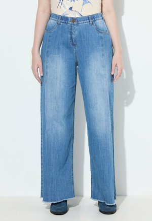 Osoba ubrana w jasnoniebieskie dżinsy o szerokich nogawkach z surowymi wykończeniami i przednimi kieszeniami, w połączeniu z niebieskimi butami i koszulą z nadrukiem ptaków.