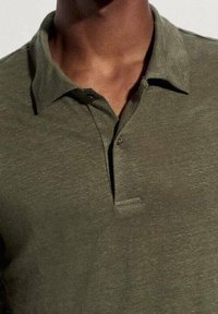 Mann in einem olivgrünen Poloshirt mit Kragen und zwei Knöpfen, vom Hals bis zur Mitte der Brust zu sehen.