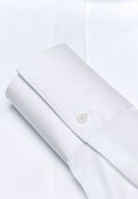 Chemise blanche en tissu lisse avec un col et un poignet arrondi. Un seul bouton est visible sur le poignet, mettant en valeur des lignes nettes.