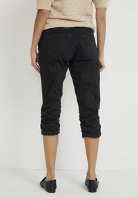 Person i svarta croppade byxor med samlade fållar, beige kortärmad stickad topp och svarta slip-on skor, stående mot en enkel vit bakgrund.