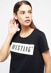 Černé tričko z bavlny s bílým obdélníkovým grafickým prvkem, na kterém je nápis "MUSTANG" tučným černým písmem. Kulatý výstřih.