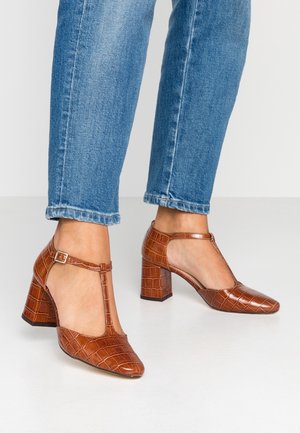 Bruine faux krokodillenleren schoenen met een T-bandontwerp en vierkante neus. Chunky hak en glanzende afwerking met verstelbare enkelgesp.