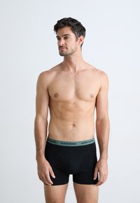 Homme torse nu avec les cheveux courts et foncés portant un boxer noir Björn Borg, se tenant contre un fond blanc uni, regardant à gauche.
