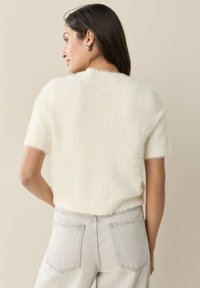 Maglione corto a maniche corte, bianco e soffice al tatto. Presenta uno scollo rotondo e una vestibilità corta, abbinato a jeans grigio chiaro.