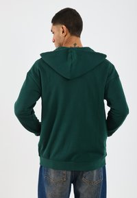 Un hoodie vert foncé avec une capuche à cordon de serrage, présentant une texture lisse et des poignets côtelés. Modèle portant un jean bleu, vu de dos.