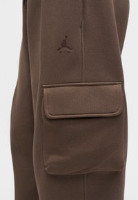 Κοντινό πλάνο καφέ παντελονιών φόρμας με μια μεγάλη πλαϊνή τσέπη με καπάκι και ένα σκούρο καφέ κεντημένο λογότυπο Jumpman Air από πάνω.