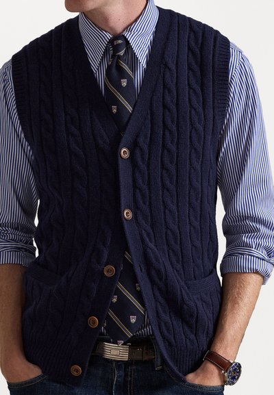 Marineblå kabelstrikket vest med v-hals, med brune knapper og framlommer, båret over en stripete blå og hvit skjorte og mønstrete slips.