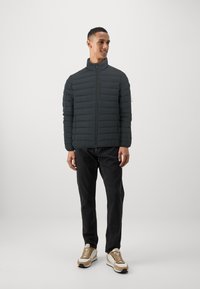 Polstret sort jakke med høj krave, lynlås lukning og horisontalt quiltet mønster, båret med mørke jeans og sneakers med beige accenter.
