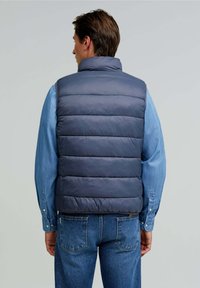Padded navy vest with high collar, featuring horizontal quilted design. Worn over a light blue denim shirt and paired with blue jeans.