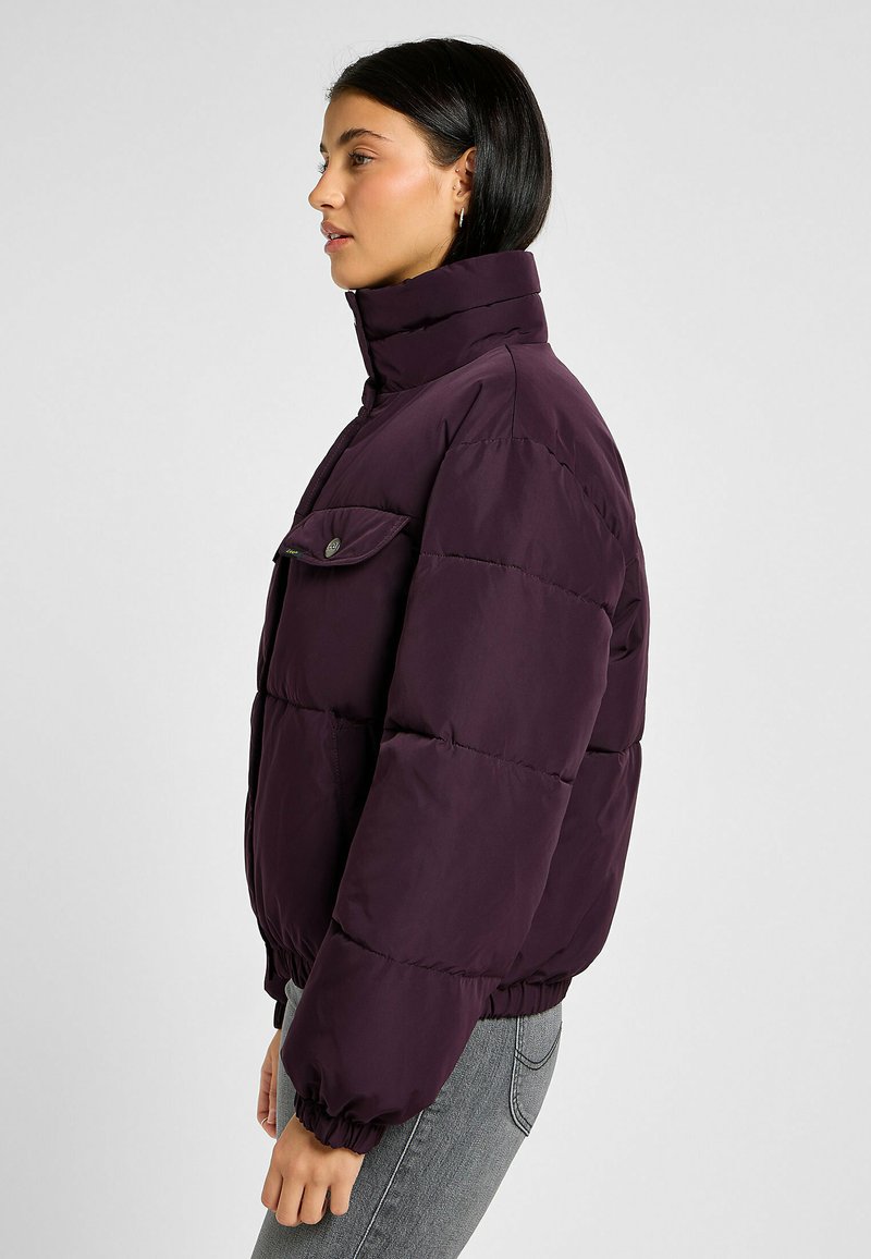 Paarse pufferjack met hoge kraag, elastische manchetten en een enkele klepzak. Voorzien van een gewatteerd ontwerp en een iets verkorte pasvorm.