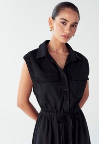 Juodas be rankovių suknelė su mygtukais priekyje, apykakle, dviem krūtinės kišenėmis ir reguliuojamu raišteliu juosmenyje. Lygus audinio paviršius.