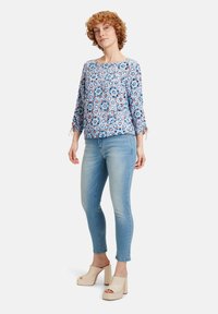 Hellblaue Bluse mit floralem Muster, 3/4-Ärmeln und Bindedetails, kombiniert mit hellblauen Skinny-Jeans und cremefarbenen Plateausandalen.