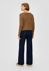 Pull en tricot marron avec col rond, associé à un jean large bleu foncé. Présente une vue de dos, une texture lisse et une palette de couleurs neutres.