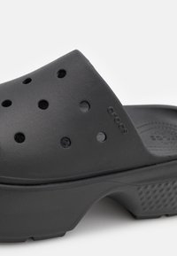 Musta Crocs-sandaalit, jotka on valmistettu kumimaisesta materiaalista, sisältävät ilmanvaihtoaukkoja, paksun kantapään ja tekstuuripohjan liukastumisen estämiseksi.