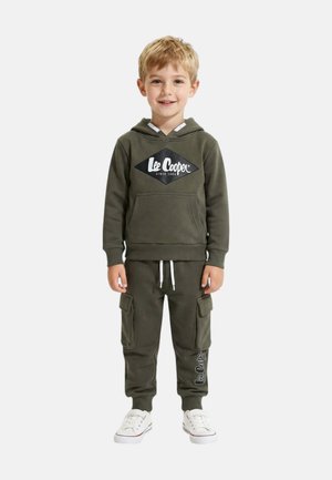 Jeune garçon souriant, portant un sweat à capuche Lee Cooper vert olive et un pantalon cargo de jogging, baskets blanches, debout devant un fond clair.