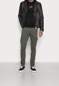 Giubbotto da biker in pelle nera sopra un hoodie nero, abbinato a pantaloni cargo verde oliva con tasche laterali e una toppa, e sneakers nere.