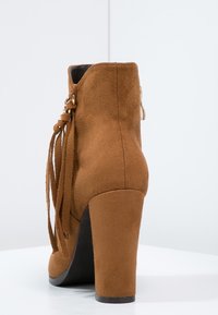 Hnědé semišové kotníkové boty na vysokém podpatku s blokovým podpatkem, bočními třásněmi a zadním zipem na bílém povrchu a pozadí.